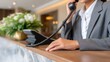 © ISVO - Female receptionist in gray suit answers phone at modern hotel front desk with floral arrangement and contemporary decor in the background
