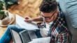 © Olya Pilipchik  - Young man remote working at home, focused on reading documents and reviewing paperwork while using a laptop for productivity in a cozy environment