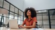 © twilight mist - Young professional woman with curly hair smiles while working on her laptop in a modern office.