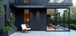 © Pete - Modern dark house exterior with wood siding, white trim, and metal balconies. Features large glass windows showing minimalist indoor furniture. Outdoor seating area has a white chair.