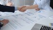 © volha_r - Business professionals shaking hands over a conference table covered with charts, graphs and financial reports, sealing a partnership and analytical, strategic agreement