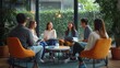 © alo - Six young adults sitting in a circle in a bright plant-filled lounge, engaged and relaxed during a collaborative meeting with laptops, notebooks and coffee on a round table
