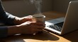 © MakarimZaydan - Pessoa com café e computador portátil em mesa sob a luz do sol.