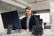 © fizkes - Portrait of smiling confident businessman in suit sits at desk in modern office, looking at camera while typing on computer keyboard. Corporate customer services, IT support, or administrative tasks