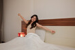 © Parichat - Young woman waking up stretching in bedroom morning
