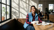 © Ozi - A smiling woman in a blue vest and plaid shirt sits at a wooden table in a sunlit cafe, holding a mug with a notebook and pen nearby.