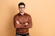 © deagreez - Confident young professional man wearing glasses with arms crossed in a brown shirt, posing against a neutral beige background for corporate portrait and business profile