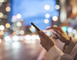 © Blada - A person's hand holds and interacts with a smartphone against a blurred, vibrant cityscape background at night, suggesting modern communication and urban life.