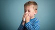 © Ava - young boy sneezing into tissue in blue striped shirt against plain background