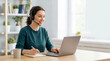 © Xenia - Smiling remote worker with headset using laptop at home. Female customer support agent or virtual assistant taking notes during online meeting