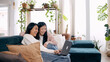 © DMegias - Two young adult women laughing together while using a laptop on a sofa in a bright living room at home