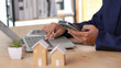 © Siwaporn - Business professional using smartphone and laptop at desk with house models symbolizing real estate investment, property management, and financial planning in a modern digital workspace environment.