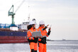 © ultramansk - Diverse industrial engineers in orange safety uniforms and hard hats collaborating with a laptop at a shipping port. Happy coworkers discussing logistics near a large cargo ship.