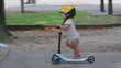 © Marco - Toddler wearing yellow helmet riding blue scooter in park pathway, captured in side profile, focused and balanced while gliding forward during playful outdoor moment