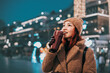 © EdNurg - Young woman smiling, enjoying coffee while walking through a beautifully lit city street at night during winter