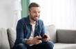 © Prostock-studio - Joyful middle aged bearded man playing video game with joystick at home, sitting on couch in living room, having fun at weekend, copy space. Home entertainment, leisure, domestic lifestyle