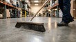 © Sri - A worker using a large industrial broom to sweep the concrete floor of a large warehouse, focusing on facility maintenance, industrial cleaning services, and professional workspace management