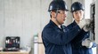 © 望見 柿元 - 日本人電気工事士の40代と20代の男性がコンクリート壁の配電盤で作業をしている屋内建設現場の様子、奥の工具箱と左側の背景余白