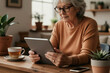 © Viktor - Elderly woman with glasses uses tablet in cozy room. She sips tea, browses internet, enjoys peaceful morning at home. Technology blends with comfort, relaxation.