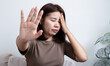 © doucefleur - Asian woman suffering from severe headache or migraine, holding temple and showing stop gesture with hand. Female feeling stressed, exhausted, and needing rest at home. Healthcare and mental health