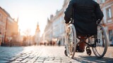 A person in a wheelchair moves along a cobblestone street in a city. Sunlight shines on the buildings and people walk nearby enjoying their time outdoors