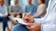 © Nataliya - Close-up of a therapist's clipboard and hand during a group session with diverse patients seated in circle, clinical group therapy concept, professional mental health documentation, diverse