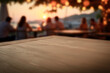 © magdal3na - Empty wooden table with clean copy space for product placement in the foreground. Blurred background with people sitting in beach outdoor bar by the sea during warm sunset. Golden light.