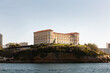 © Simplementbeau. - Palais du Pharo à Marseille vue depuis la mer, rocher avec de la verdure en contre-bas, mer au premier plan, lumière douce et ensoleillée, ciel bleu
