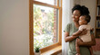 © Xenia - Smiling African American mother holding her baby and looking out the window. Happy black woman with infant child at home. Family love and motherhood concept
