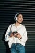 © BullRun - Cheerful young woman with braces using mobile phone in headphones and holding takeaway coffee. Girl enjoying digital connectivity, streaming music or chatting in contemporary urban setting.