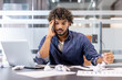 © Tetiana - Thoughtful young Indian man sitting at a desk in the office, holding his head in his hands and looking at the crumpled paper in frustration