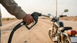 © ng-TDRD - A person refuels a black motorcycle on a dirt road in a desert-like area, holding a fuel pump nozzle close to the bike's tank.