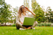 © maxbelchenko - A happy woman sits on the grass in a park with a laptop on a sunny day. A female freelancer works on a laptop in a park at sunset. Concepts for remote work, education, or leisure.