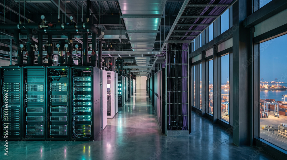 Server racks line the walls of a large data center. It is dusk, and the ...