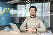 © Liubomir - Smiling bearded professional wearing glasses sits at a modern desk with laptop and office supplies, looking confident and relaxed while working remotely in a contemporary workspace