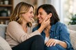 © by.anastasiya - Two adult women sit close together on a sofa, smiling and laughing while one touches the other’s face in a bright living room.