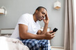 © Anastasiya - African american man sitting on bed looking at smartphone with worried expression in bedroom. Stressful lifestyle moment at home, communication, problem solving and anxiety concept