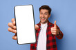 © Prostock-studio - A man stands in front of a blue background, holding a blank smartphone screen in one hand while giving a thumbs up with the other hand. He appears happy and engaged in the moment.