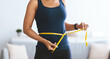 © Prostock-studio - A woman stands indoors and uses a yellow measuring tape to measure her waist. She focuses on her body measurements as part of her fitness routine. The setting is bright and open.