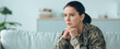 © Prostock-studio - A soldier in military attire sits on a couch with hands clasped under her chin. She appears deep in thought in a cozy living room. The room has soft lighting and simple decor.