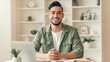 © Prostock-studio - A young man sits at a desk with a laptop, smiling at the camera. Shelves in the background hold plants and decorative items. The room has good lighting and a modern feel.