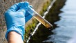 © Netsai - A gloved hand holds a test tube containing a murky water sample with algae, next to a body of water.