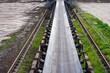 © Adam - Long industrial rubber conveyor belt system with metal idler rollers, extending into the distance parallel to heavy machine travel rails at an open-pit mining or industrial facility.