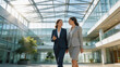 © Ricky - Two professional businesswomen in formal suits walking and talking together in a modern, sunlit corporate office atrium with glass and steel architecture and bright natural light streaming in.