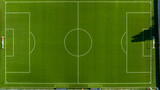 Perpendicular top-down aerial view of a vibrant green synthetic soccer field, showcasing precise white boundary lines, the center circle, penalty areas, and goals under clear, bright daylight.