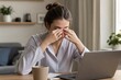 © Studio - Stressed young professional woman rubbing eyes while working remotely on laptop at home, exhausted from long hours in cozy indoor setting with warm lighting and coffee mug