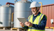 © NAD SANG PAP - Mature male worker in safety helmet using tablet at industrial plant, smiling with high visibility vest and tanks in background
