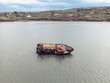 © AmazingAerialAgency - Aerial view of a rusty shipwreck abandoned in shallow coastal waters with green hills and houses in the background under an overcast sky.