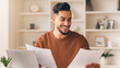 © Prostock-studio - A man sits at a desk with a laptop in front of him. He holds papers in both hands and looks pleased while reading. The room has shelves and a plant nearby, suggesting a home office environment.