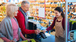 © Prostock-studio - A man and a woman stand at the checkout counter in a grocery store. The cashier helps them complete their purchase. The store is filled with various food items on the shelves.